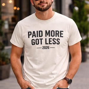 Puede incluir: Camiseta color crema con el texto negro "PAID MORE GOT LESS - 2026". La persona lleva gafas de sol y una gorra de béisbol.