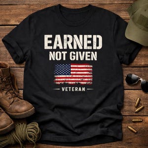 May include: Black t-shirt with the text "EARNED NOT GIVEN" above a distressed American flag graphic. Below the flag, the word "VETERAN" is printed. The shirt is displayed on a wooden surface with boots and accessories.
