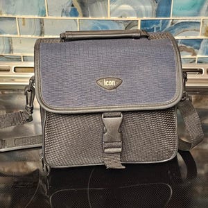 May include: A navy blue and black messenger bag featuring a textured fabric flap with a centered oval patch displaying the word icon. The bag includes a top handle, an adjustable shoulder strap, and a central black plastic buckle closure.