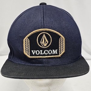 May include: A navy blue baseball cap with a flat black brim. The front features a black patch with a gold embroidered border, a gold Volcom stone logo, and the word VOLCOM in white text. The patch has decorative gold side stitching details.