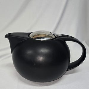 May include: A matte black teapot with a rounded body and a curved handle. The teapot has a silver-colored lid and spout. The design is modern and minimalist, suitable for brewing tea.