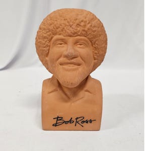 May include: A terracotta-colored ceramic bust of Bob Ross featuring his signature curly afro hairstyle and beard. The sculpture depicts the head and shoulders of the artist with the name Bob Ross printed in black script on the front base.
