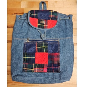 May include: Blue denim backpack with a patchwork design. The backpack features a front zippered pocket and a flap with a red and plaid pattern. The top flap has a small metal emblem. The bag has a drawstring closure and a top handle.