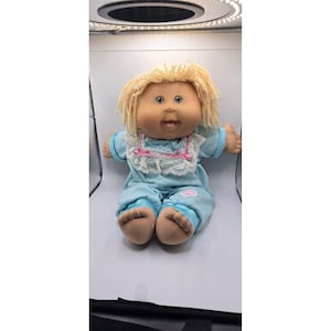 May include: A vintage Cabbage Patch Kid doll with blonde hair and blue eyes. The doll is wearing a light blue onesie with a white lace collar and pink accents. The doll is sitting with its arms outstretched.