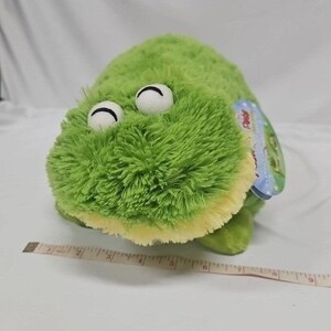 May include: A plush green frog toy with white eyes and a yellow belly. The frog has a tag attached to its side. A measuring tape is next to the frog.