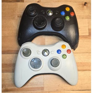 May include: Two video game controllers, one black and one white, featuring a central Xbox guide button, two analog sticks, a directional pad, and four colored action buttons labeled A, B, X, and Y, along with Back and Start buttons.