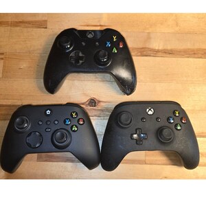 May include: Three black video game controllers arranged in a triangular formation. Each controller features two analog sticks, a directional pad, and four action buttons labeled with the letters A, B, X, and Y in green, red, blue, and yellow colors.
