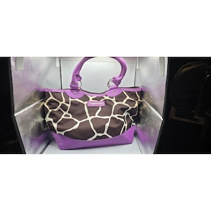 May include: A Tupperware tote bag with a giraffe print pattern in brown and white. The bag has a purple base, trim, and handles. The handles are arched and connected to the bag with silver rings. The Tupperware logo is visible.