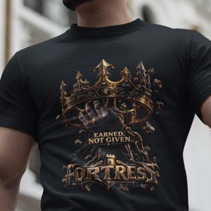 Può includere: Una t-shirt nera a maniche corte con una stampa grafica di una corona d'oro metallico tenuta da una mano. Sotto la corona, il testo recita "EARNED, NOT GIVEN." e "FORTRESS" in un carattere dorato stilizzato con piccoli frammenti.