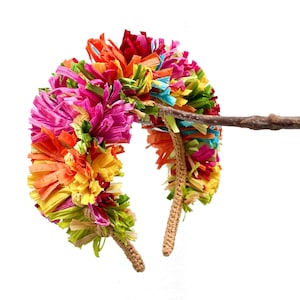 May include: A headband featuring a dense arrangement of multi-coloured raffia strips in shades of hot pink, bright orange, sunny yellow, lime green, and turquoise blue. The base of the headband is covered in a tan, crocheted or woven material.