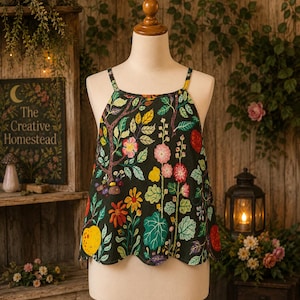 May include: A sleeveless black top featuring a colorful botanical print with various flowers, leaves, and fruits. The garment has thin shoulder straps, a straight neckline, and a scalloped hemline, displayed on a mannequin torso.