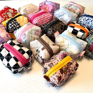 May include: A collection of small, quilted fabric pouches in various colours and patterns, including checkered, floral, and geometric prints. Each rectangular pouch features a top zip closure in a contrasting colour and a soft, padded texture.