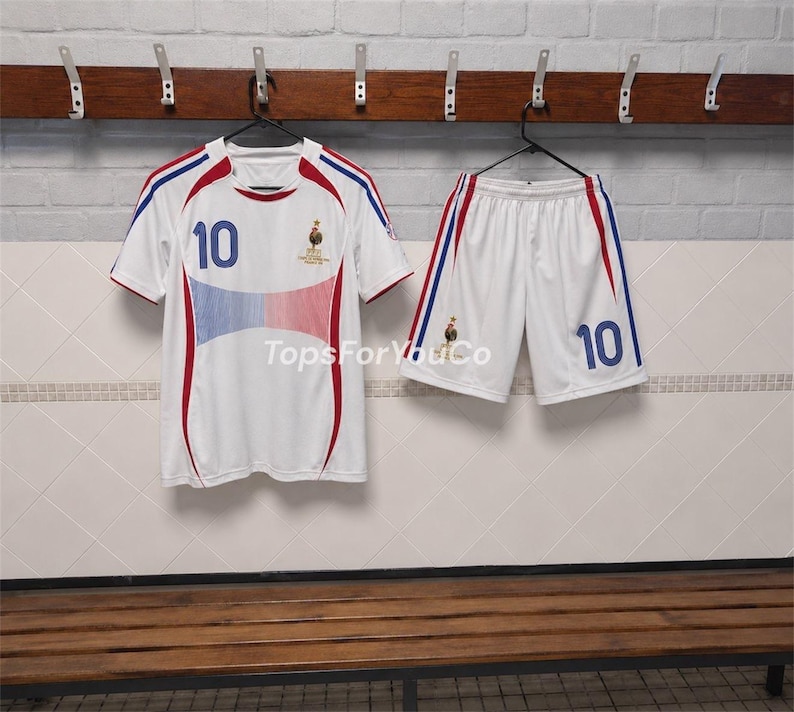 Puede incluir: Un conjunto de f&uacute;tbol blanco que incluye una camiseta y pantalones cortos con detalles en azul y rojo. La camiseta muestra el n&uacute;mero 10 en azul, un emblema de gallo y el texto TopsForYouCo. El pantal&oacute;n corto tambi&eacute;n tiene el n&uacute;mero 10 y un gallo.
