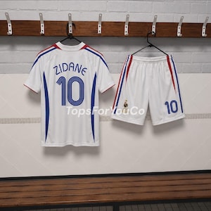 Puede incluir: Un conjunto de camiseta y pantal&oacute;n corto de f&uacute;tbol blanco con detalles en azul y rojo. La camiseta muestra el nombre ZIDANE y el n&uacute;mero 10 en azul. El pantal&oacute;n corto presenta el n&uacute;mero 10 y un peque&ntilde;o emblema. El texto TopsForYouCo es visible.