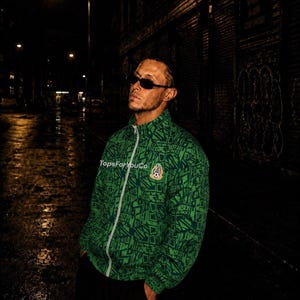 May include: A green zip-up track jacket featuring a repeating geometric pattern and the word Mexico printed throughout. The jacket includes a central white zipper and a circular emblem on the chest with a soccer ball graphic and text TopsForYouCo.