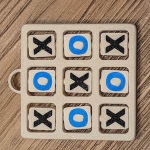May include: A 3D-printed tic-tac-toe game with a light beige frame. The game pieces are white with blue circles and black X's. The game board has a small handle on the side.