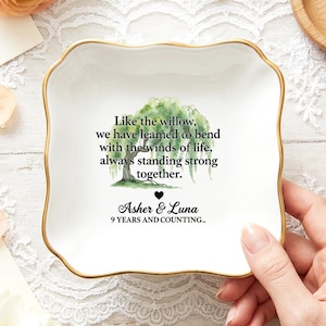 Op de afbeelding: Een vierkant wit keramieken schaaltje met een goudkleurige, geschulpte rand. Het midden bevat een aquarelillustratie van een wilg, de tekst "Like the willow, we have learned to bend with the winds of life, always standing strong together.", een hartje en de namen "Asher & Luna" met "9 YEARS AND COUNTING.." eronder.