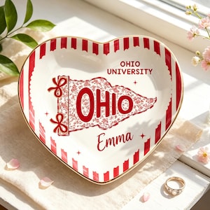 May include: Heart-shaped ceramic dish with a red and white striped border and gold trim. The center features a red floral design with the word "OHIO" and "OHIO UNIVERSITY" in red. The name "Emma" is also printed on the dish.