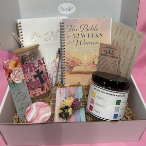 May include: A gift box filled with inspirational items. Includes a journal, a Bible study book, a glass with "PRAY" text, a candle, a pen, a bookmark, and a small floral arrangement. The box is white with a pink interior.
