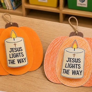 May include: Two pumpkin-shaped hanging decorations made of paper. One is solid orange, the other is coloured with orange crayon. Each features a central candle illustration with a flame and the text 'JESUS LIGHTS THE WAY' with a small cross symbol.