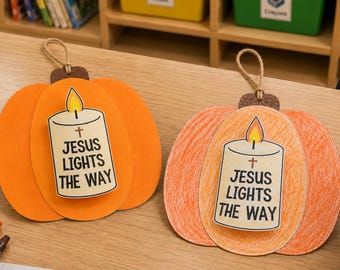 May include: Two pumpkin-shaped hanging decorations made of paper. One is solid orange, the other is colored with orange crayon. Each features a central candle illustration with a flame and the text 'JESUS LIGHTS THE WAY' with a small cross symbol.