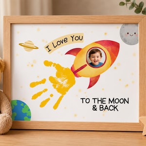May include: A framed print with a child's photo in a rocket ship illustration. The print includes a handprint, a planet, a smiling moon, and the text "I Love You To The Moon & Back". The frame is made of light brown wood.
