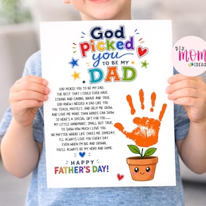 May include: A child holds a Father's Day print with the text "God picked you to be my Dad". It includes a handprint and a cartoon plant pot. The words "Happy Father's Day!" are also visible. The print is in full color.