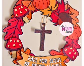 May include: A decorative wreath featuring paper cutouts of red and orange autumn leaves, pumpkins, and red mushrooms with white spots. A brown cross hangs in the center from a gold string. An orange banner at the bottom reads FALL FOR JESUS HE NEVER LEAVES.