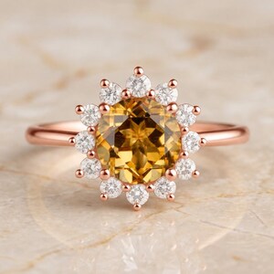 May include: A rose gold ring featuring a large, round, golden-yellow center stone surrounded by a halo of twelve small, clear, round-cut stones. The band is thin and polished, with the entire setting crafted in a warm, metallic rose gold tone.