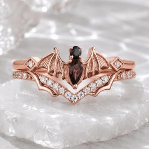 May include: A rose gold ring set featuring a bat-shaped centrepiece with a black round stone head and a marquise-cut dark red stone body. The wings are detailed with textured lines, and the band is accented with small clear stones in a V-shape.