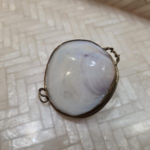 May include: A white seashell with subtle purple hues, encased in a gold-toned metal frame. The shell has a smooth, glossy surface and is set against a neutral background. The frame has decorative, looped handles.