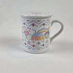 May include: A white ceramic mug with a matching lid, featuring a colorful cross-stitch pattern of a small house, a rainbow, a heart, and geometric floral motifs in shades of blue, yellow, red, and purple, complete with a curved handle.