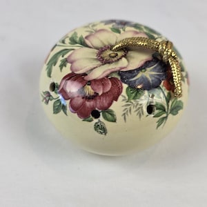 以下が含まれることがあります： ピンク、紫、白の花と緑の葉が描かれた、クリーム色の丸いセラミック製ポマンダー。本体には小さな円形の通気孔があり、上部には金色の金属製コードループが付いています。