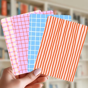 Könnte beinhalten: Eine Hand hält vier rechteckige Karten mit bunten Mustern. Die Muster umfassen ein rosa-oranges Karomuster, ein rosa Gitter, ein blaues Gitter und orange-weiß gestreifte Vertikalstreifen. Im Hintergrund ist ein verschwommenes Bücherregal zu sehen.