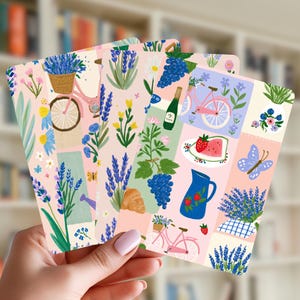 Pode incluir: Um conjunto de cartões retangulares com uma colagem de ilustrações, incluindo ramos de alfazema, bicicletas cor-de-rosa, um jarro azul com morangos, uma fatia de melancia, um croissant, uma garrafa de vinho, uma borboleta e motivos florais.