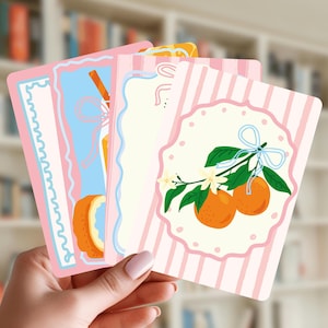 May include: A set of rectangular cards featuring pastel designs. One card displays two oranges on a branch with white blossoms and a light blue ribbon, set against a pink and white striped background with a scalloped circular frame.