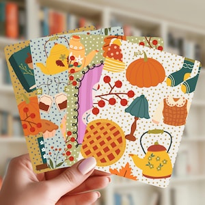 May include: A set of four rectangular cards featuring a repeating pattern of autumn-themed illustrations including orange pumpkins, yellow teapots, red berries, knitted hats, socks, a lamp, and a lattice-topped pie on a cream background with brown dots.