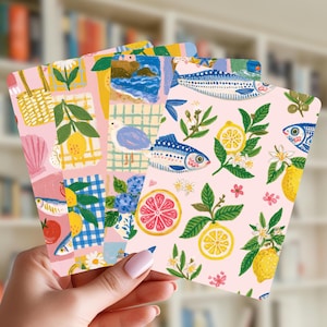 May include: Three rectangular cards featuring colourful illustrations on a light pink background. The designs include lemons, citrus slices, green leaves, white blossoms, blue fish, a seagull, a beach scene, a woven bag, and a blue checkered pattern.