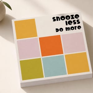May include: A square notepad featuring a grid of colorful squares in yellow, light pink, orange, olive green, and light blue. The text "snooze less do more" is printed in black, rounded, lowercase letters in the upper right corner of the page.