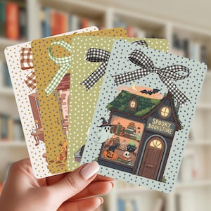 May include: A set of four rectangular cards featuring a polka dot background and a gingham ribbon at the top. Each card displays a different illustration of a building, including one labeled Spooky Bookstore with a ghost, cat, and pumpkin.