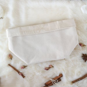 May include: A cream-colored canvas pouch with a folded-over top. The pouch is rectangular with a slightly tapered bottom. The background is a white, fluffy surface with cinnamon sticks, dried rosebuds, and star anise.