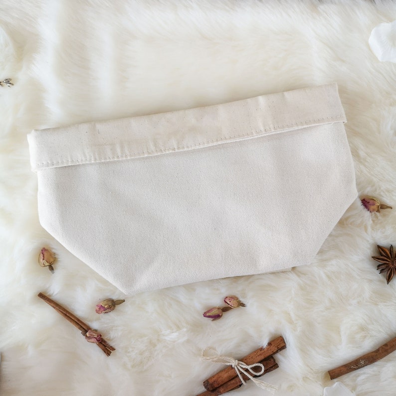 May include: A cream-colored canvas pouch with a folded-over top. The pouch is rectangular with a slightly tapered bottom. The background is a white, fluffy surface with cinnamon sticks, dried rosebuds, and star anise.