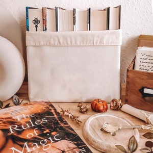 May include: A cream-colored canvas storage bin filled with books. The book spines are visible above the bin's edge. A copy of "The Rules of Magic" is in the foreground, along with decorative items like a small pumpkin and a wooden spoon.