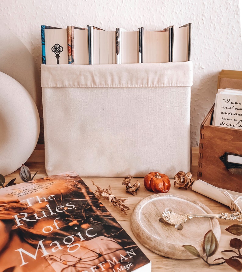 May include: A cream-colored canvas storage bin filled with books. The book spines are visible above the bin's edge. A copy of "The Rules of Magic" is in the foreground, along with decorative items like a small pumpkin and a wooden spoon.