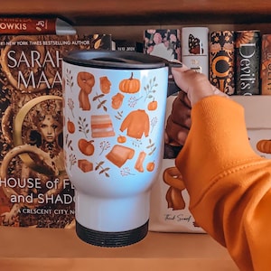 May include: White travel mug with a black lid, featuring an autumn-themed design. The design includes orange pumpkins, sweaters, scarves, and leaves. The mug is held in front of a bookshelf with books.