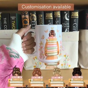 May include: White ceramic mug featuring a whimsical illustration of a person carrying a stack of books, with butterflies and floral accents. The mug has the text "Customisation available". The mug is held by a person.