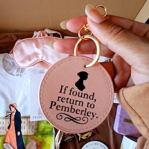 May include: A round, blush-colored keychain with gold hardware. The keychain features the silhouette of a woman and the text "If found, return to Pemberley." Other items in the image include a sleep mask and a book.