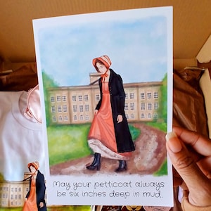 May include: A watercolour print depicting a woman in a coral dress and black coat, walking on a path. The text reads, "May your petticoat always be six inches deep in mud." The background shows a large building and green trees.