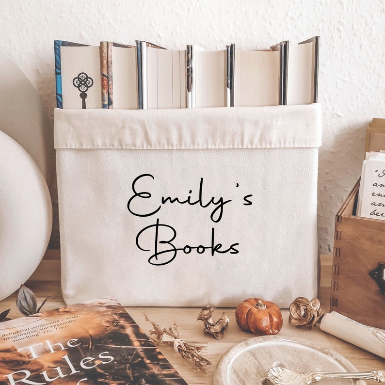 May include: A cream-colored canvas book bin with the text "Emily's Books" in black script. The bin is filled with books and sits on a wooden surface. Decorative items like a small pumpkin and a bundle of dried herbs are nearby.