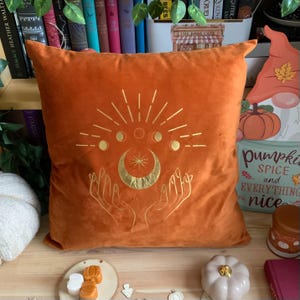 May include: Orange velvet cushion with gold embroidery of a crescent moon, stars, and hands. The cushion has the text "pumpkin spice and everything nice" on the side.